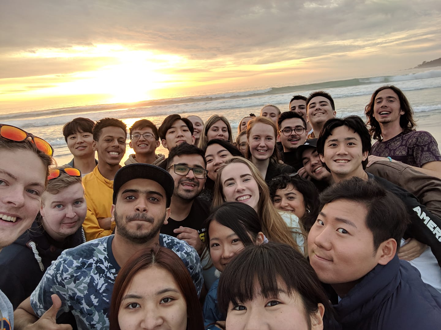 A crowd of enthusiastically smiling I-House students posing for a group selfie with a California sunset on the Pacific Ocean behind them.