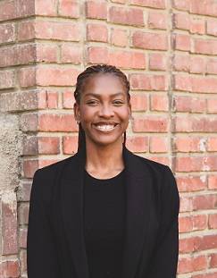 Image of young woman smiling in front of a brick building. She has deep brown skin tone, braids, and a black shirt and blazer.
