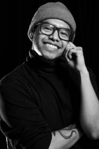 A black-and-white portrait of a smiling person wearing a beanie, glasses, and a turtleneck sweater. They have a septum piercing and an animal tattoo on their forearm. Their pose is relaxed, with one hand resting on their face, and they are looking directly at the camera. The background is dark, creating a contrast that highlights their facial features and expression.